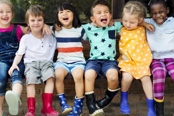 A group of toddler children of all different ethnicities sit side by side with their arms around each other while smiling